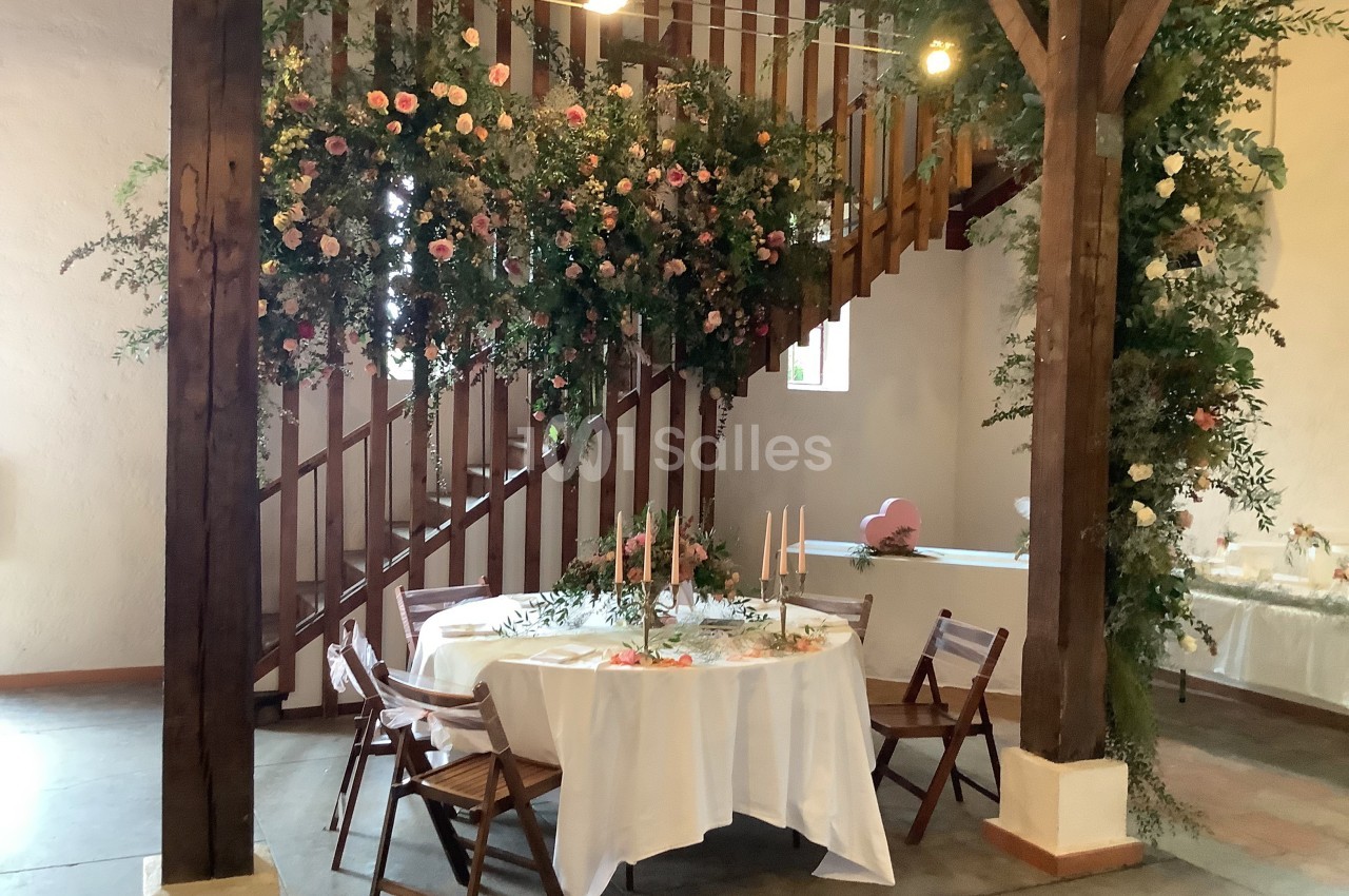 Table ronde décorée de bougies et fleurs sous un escalier en bois, avec une suspension florale et chaises en bois.