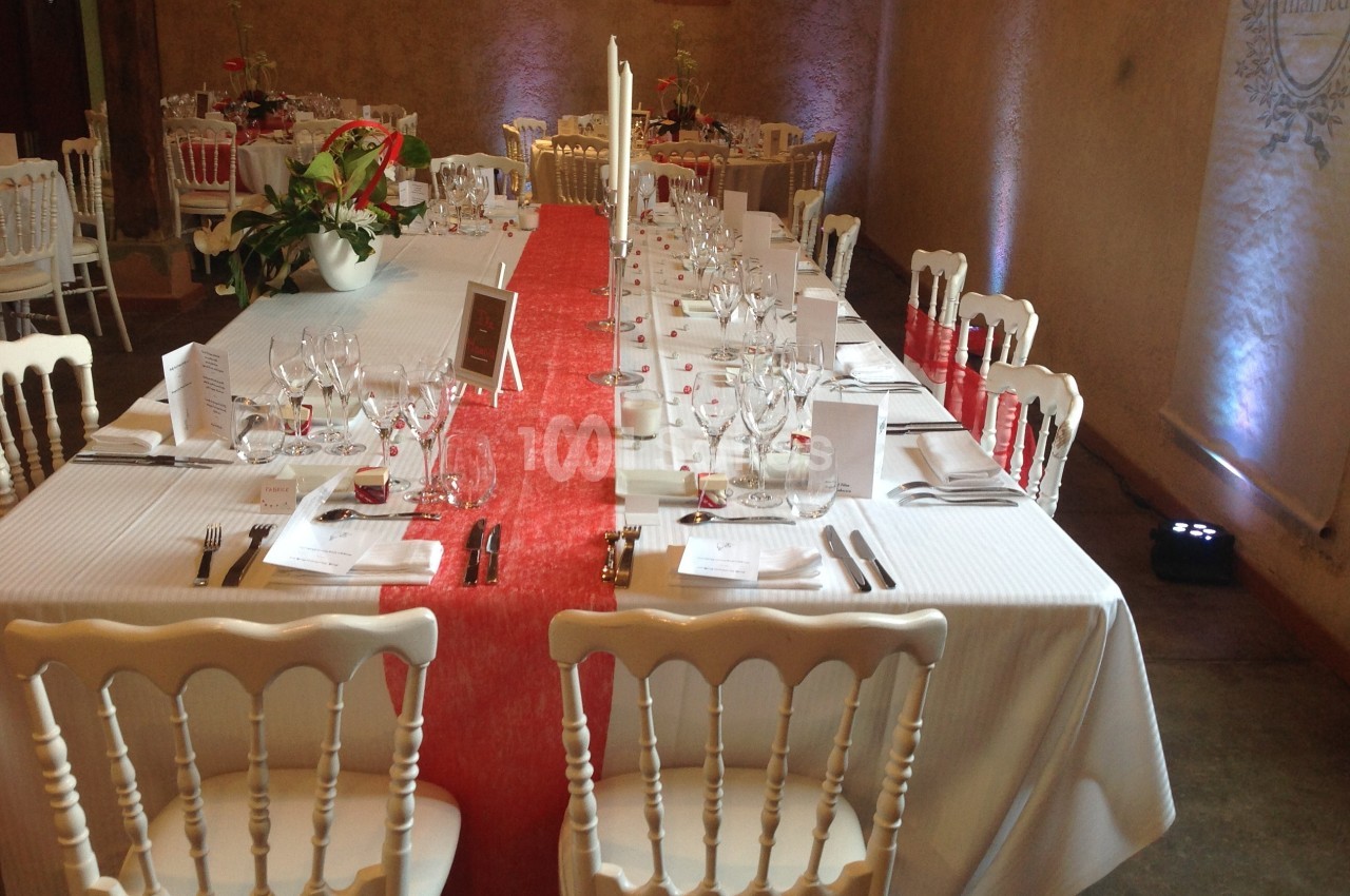 Table de réception élégamment dressée avec nappes blanches, chemin de table rouge et chaises en bois blanc.
