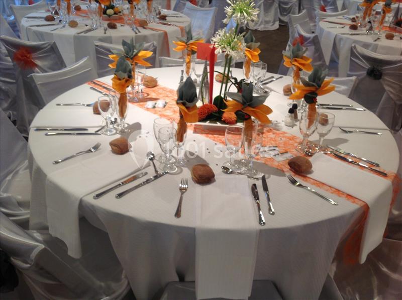 Table ronde dressée pour un événement, avec nappes blanches, couverts disposés et décorations florales orange.