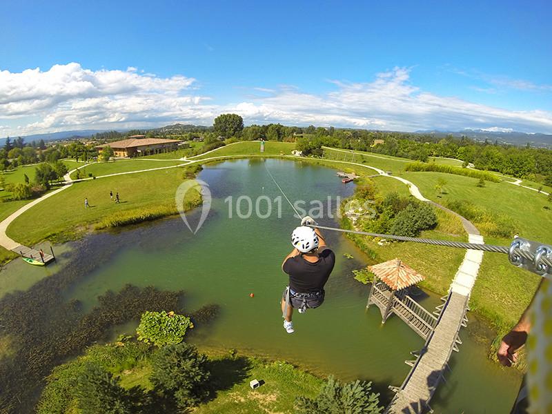 Une personne en tyrolienne traverse un lac entouré de verdure et de sentiers sous un ciel dégagé.