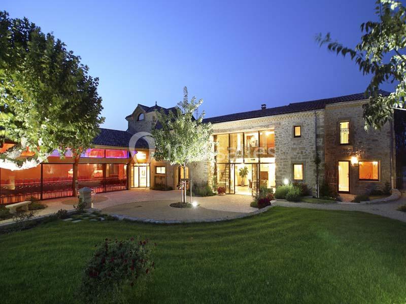 Bâtisse en pierre éclairée de nuit, entourée d'un jardin avec pelouse, arbres et terrasse vitrée illuminée.