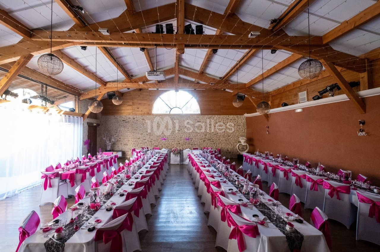 Salle de réception décorée pour un mariage, avec des tables alignées et nappes blanches ornées de nœuds roses.