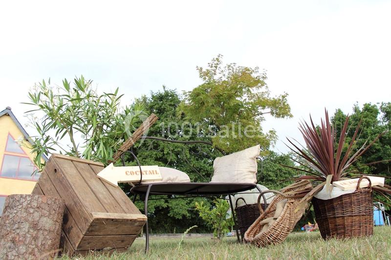 Mobilier de jardin avec coussins, paniers en osier, plantes et pancarte ’cocktail’ dans un cadre extérieur verdoyant.