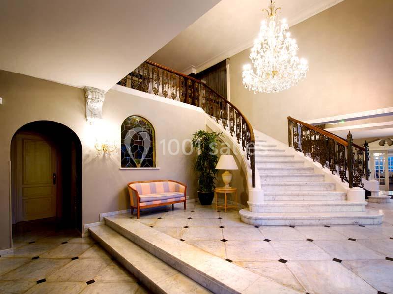 Escalier en marbre avec rampe en fer forgé dans un hall élégant, éclairé par un grand lustre.