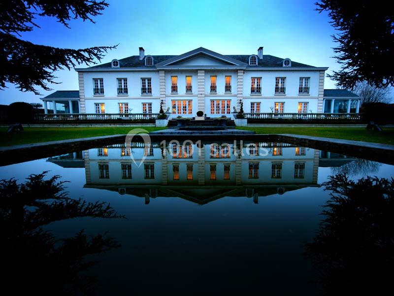Façade d'un grand bâtiment blanc éclairé, reflété dans un bassin au premier plan, entouré de végétation.