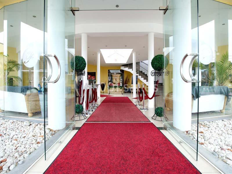 Entrée d'un bâtiment avec tapis rouge, portes vitrées ouvertes, colonnes blanches et décoration végétale.