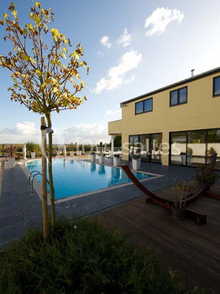 Maison moderne avec piscine extérieure, terrasse en bois, chaises longues et ciel dégagé en arrière-plan.