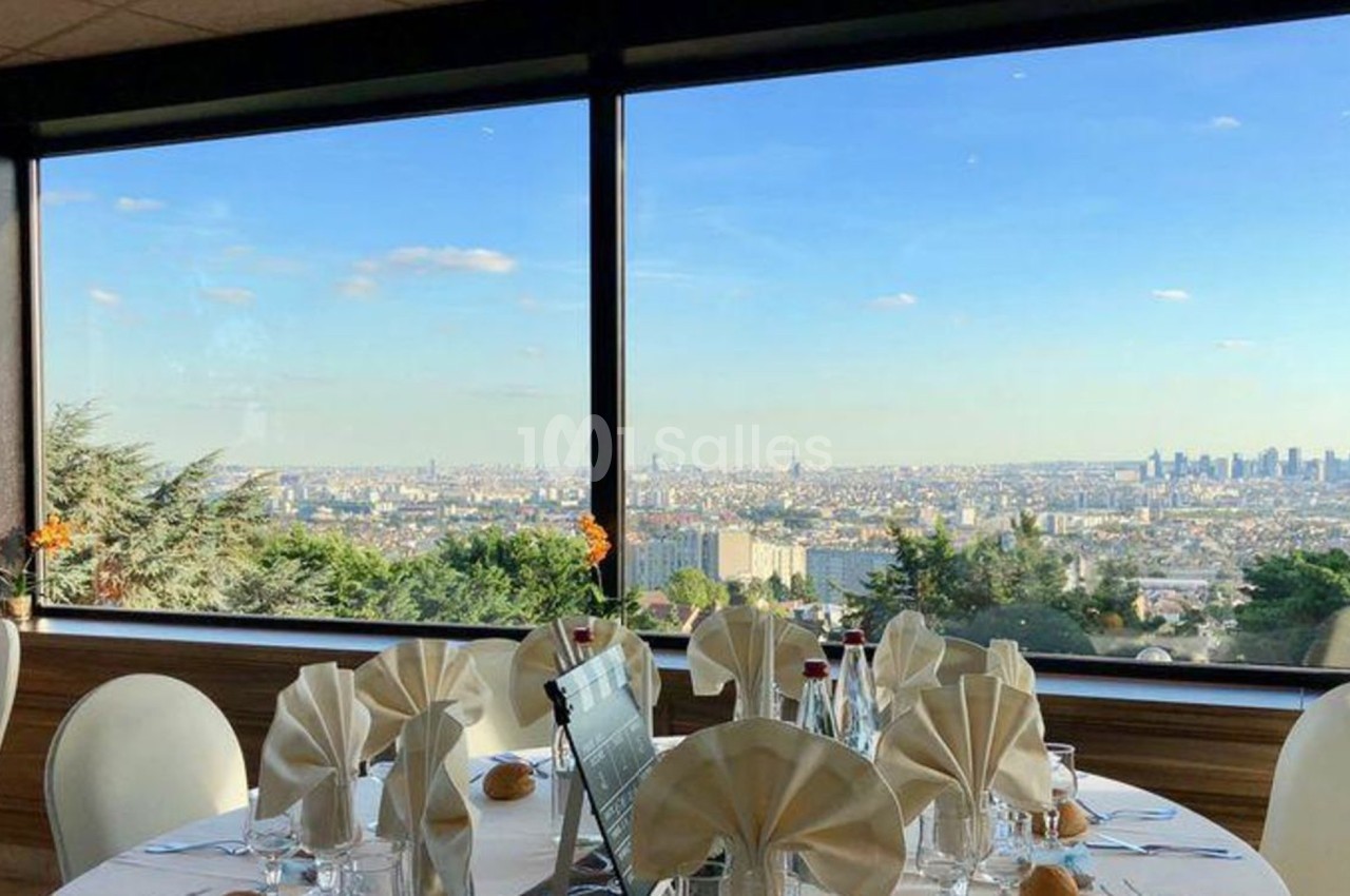 Vue panoramique sur une ville depuis une salle de réception avec des tables dressées et des grandes baies vitrées.