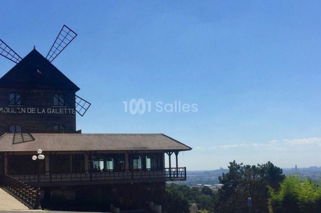 Moulin en bois avec des ailes, inscrit ’Moulin de la Galette’, surplombant un paysage urbain sous un ciel dégagé.