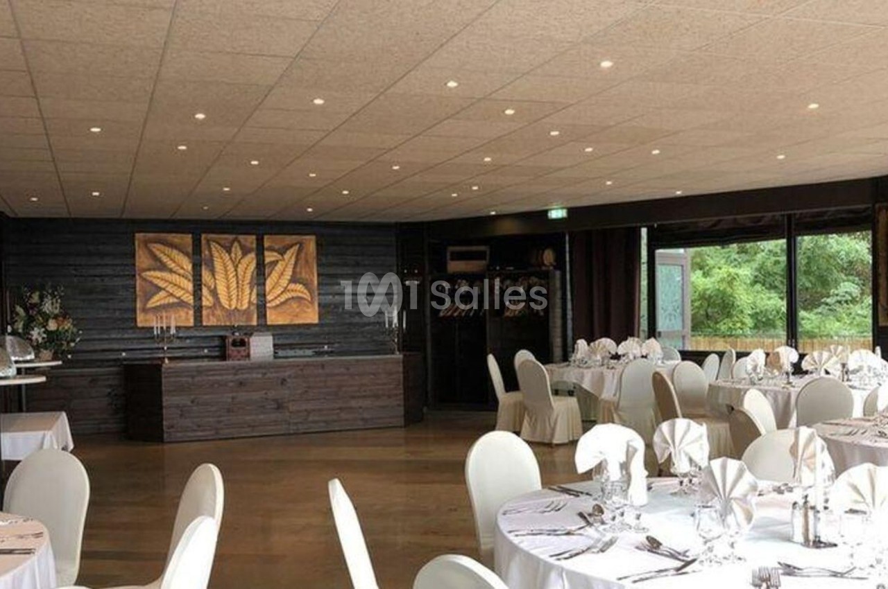 Salle de réception lumineuse avec tables rondes dressées, chaises blanches et grandes baies vitrées donnant sur un espace…