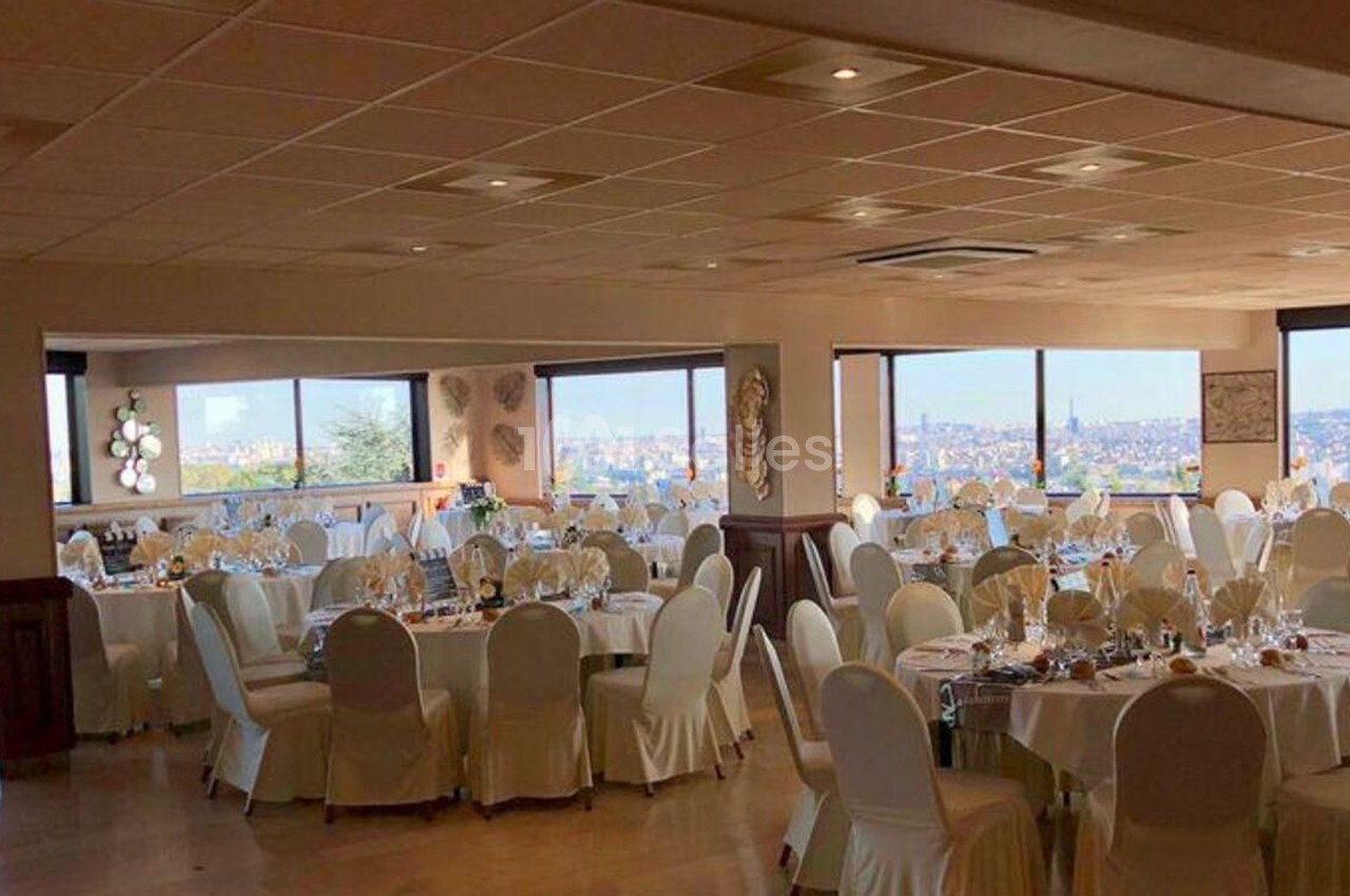 Salle de réception lumineuse avec tables rondes dressées, nappes blanches et vue panoramique sur une ville.