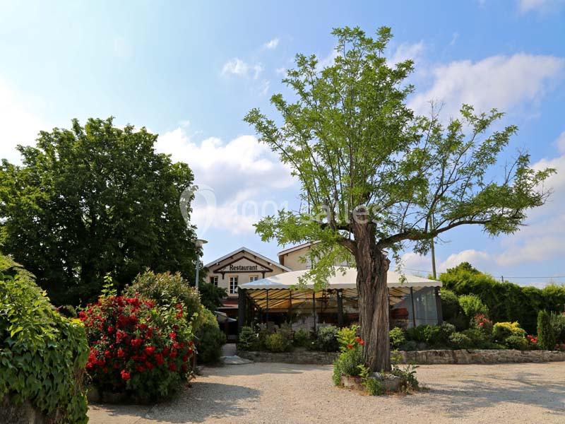 Cour extérieure d'un restaurant avec terrasse ombragée, arbres et arbustes fleuris sous un ciel partiellement nuageux.