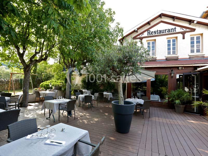 Terrasse en bois d'un restaurant avec tables dressées, chaises en osier, arbres et façade en arrière-plan.