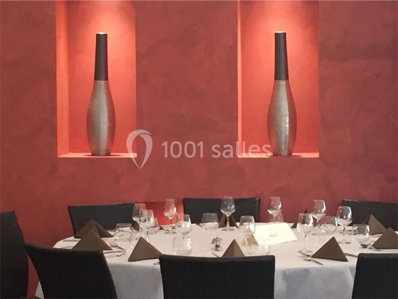 Table ronde dressée avec nappes blanches, serviettes pliées et verres, devant un mur rouge avec niches décoratives.