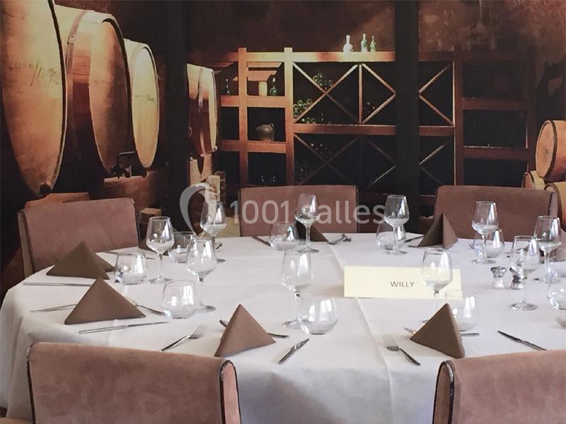 Table dressée avec nappes blanches, serviettes pliées et verres, dans une salle décorée de tonneaux et étagères à vin.