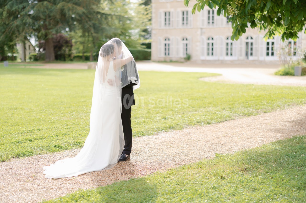 Un couple de mariés s'embrasse sous un voile dans un jardin, près d'un bâtiment en pierre avec de grandes fenêtres.