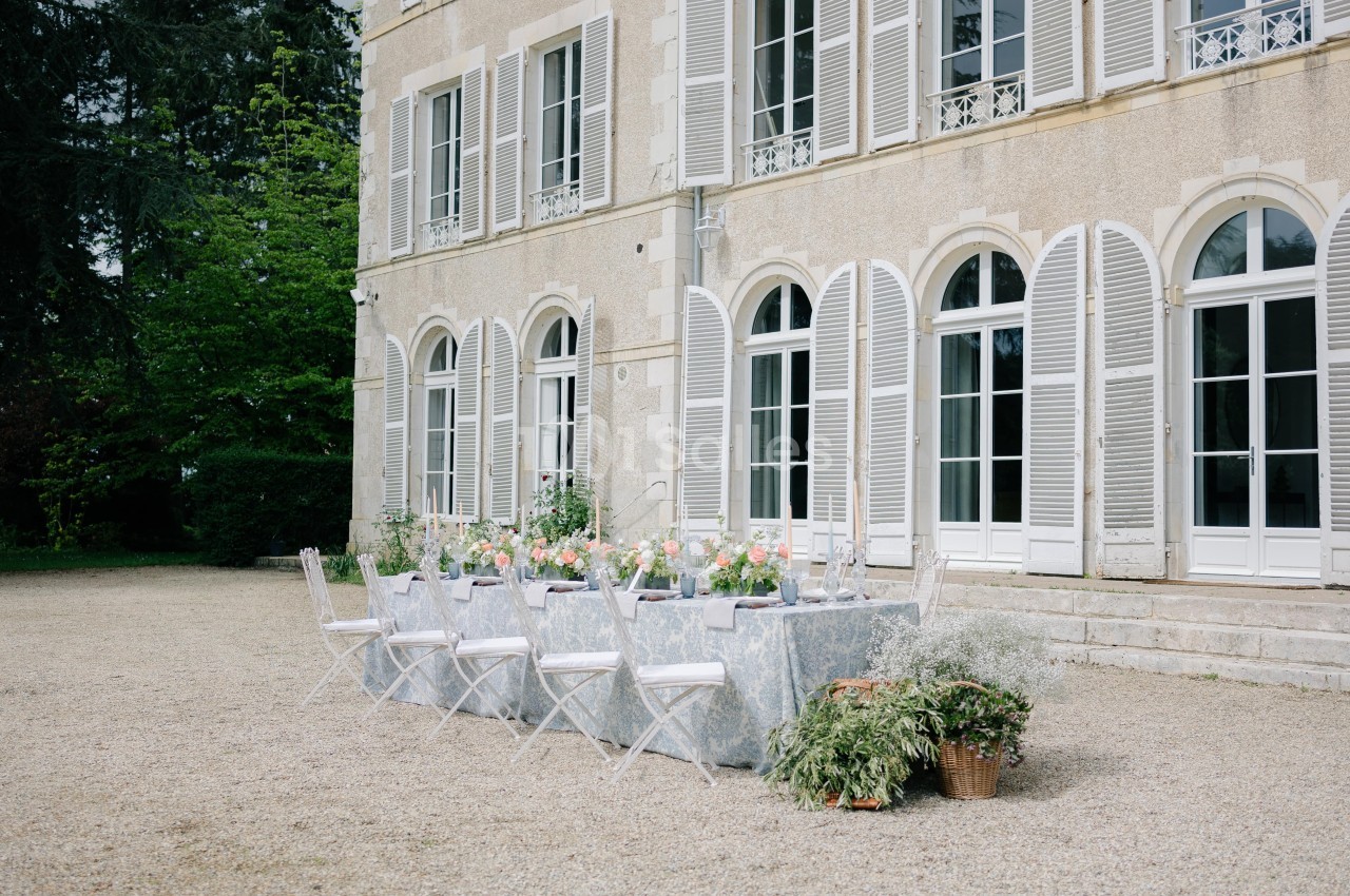 Table dressée en extérieur avec des chaises blanches devant une grande bâtisse aux volets ouverts.