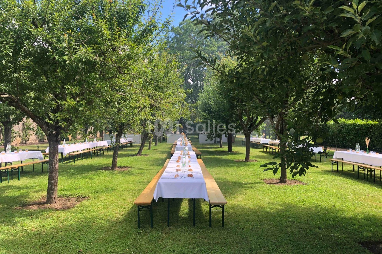 Tables alignées avec nappes blanches et bancs en bois disposés dans un jardin verdoyant sous des arbres.