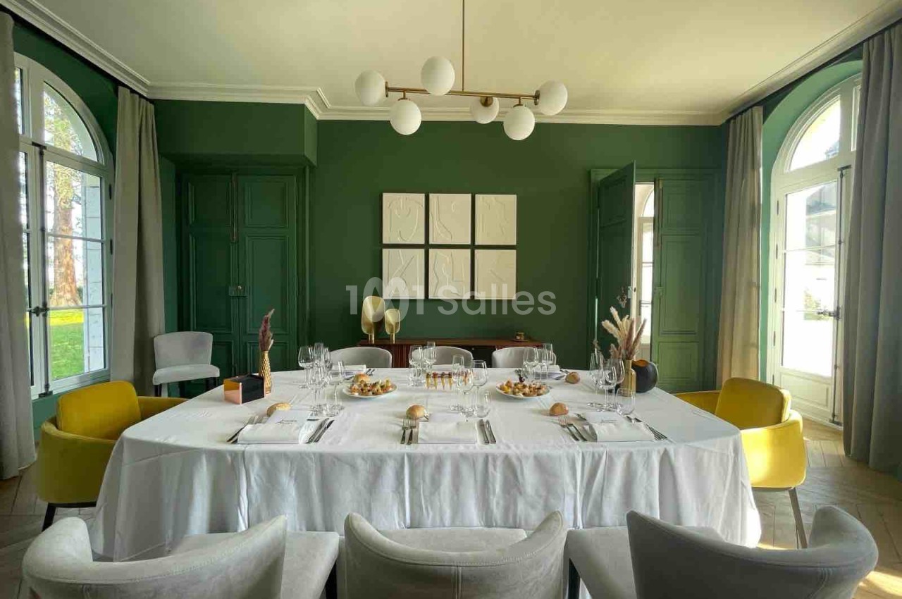 Salle à manger élégante avec murs verts, grande table dressée avec nappes blanches, chaises variées et lumière naturelle.