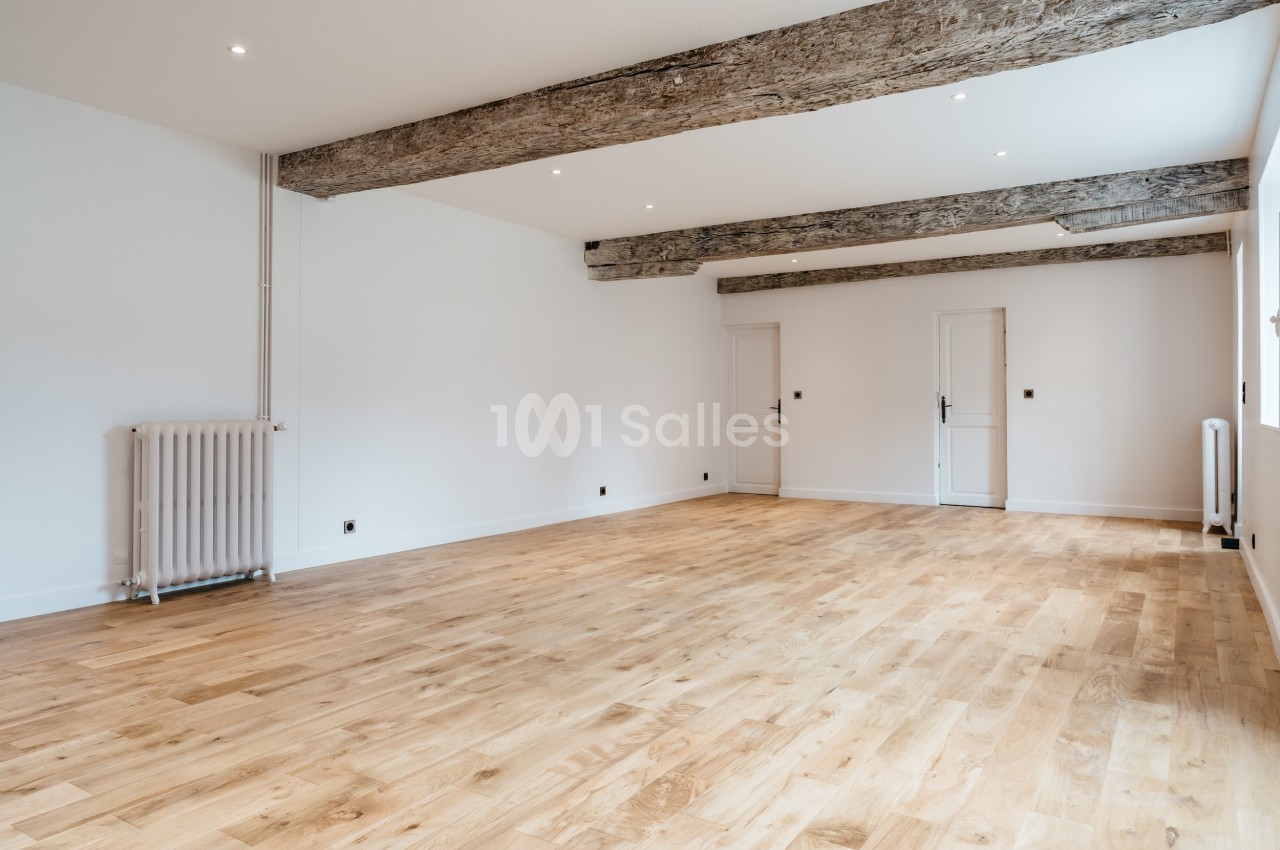 Salle vide avec parquet en bois clair, murs blancs, poutres apparentes au plafond et radiateurs en fonte.