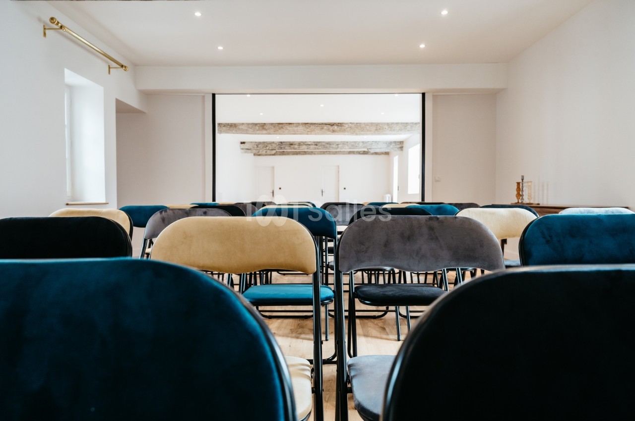 Chaises alignées dans une salle lumineuse avec un plafond en poutres apparentes et un écran à l'arrière.