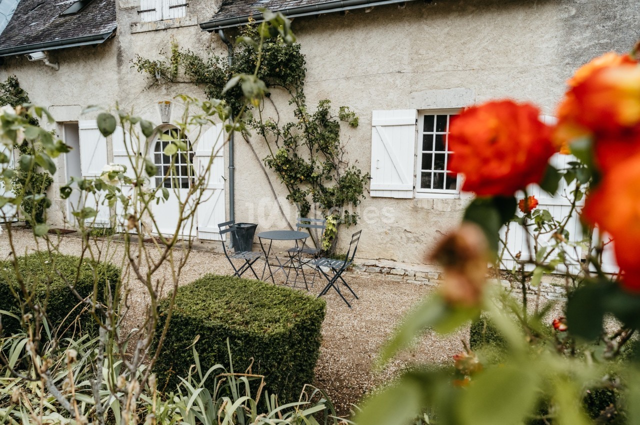 Façade d'une maison en pierre avec volets blancs, table et chaises en métal dans une cour fleurie.