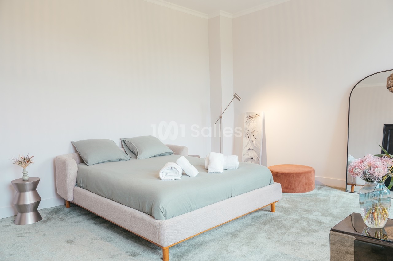 Chambre lumineuse avec un lit double, linge de maison plié, miroir, lampe sur pied et décoration florale.
