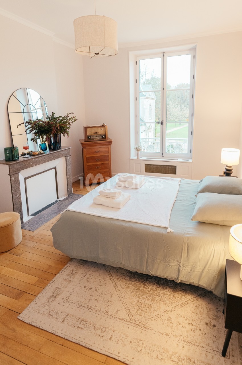 Chambre lumineuse avec lit double, parquet, cheminée décorative, miroir, et vue sur un jardin par une grande fenêtre.