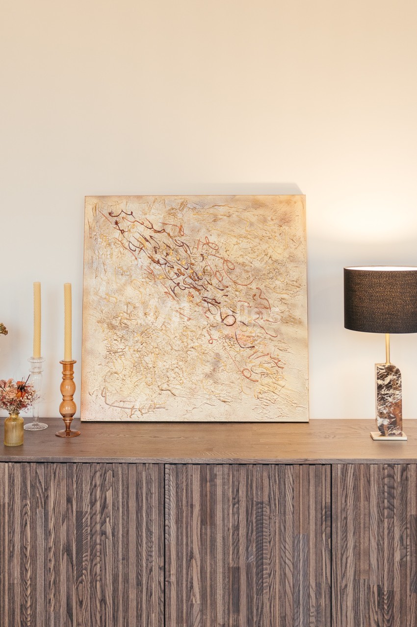 Peinture abstraite beige et dorée accrochée au-dessus d'un buffet en bois, décoré de bougies, fleurs et lampe moderne.