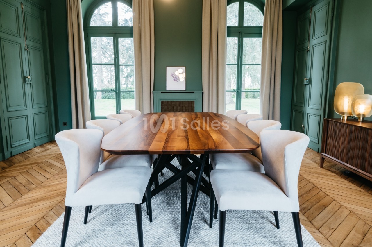 Salle à manger avec table en bois, chaises rembourrées claires, parquet en chevrons et grandes fenêtres avec rideaux beiges.
