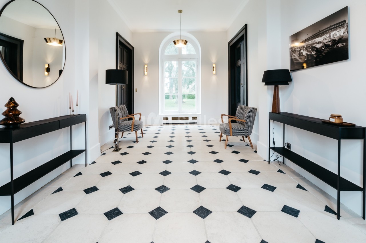Hall lumineux avec sol en carreaux noirs et blancs, deux fauteuils, tables d'appoint et grande fenêtre centrale.
