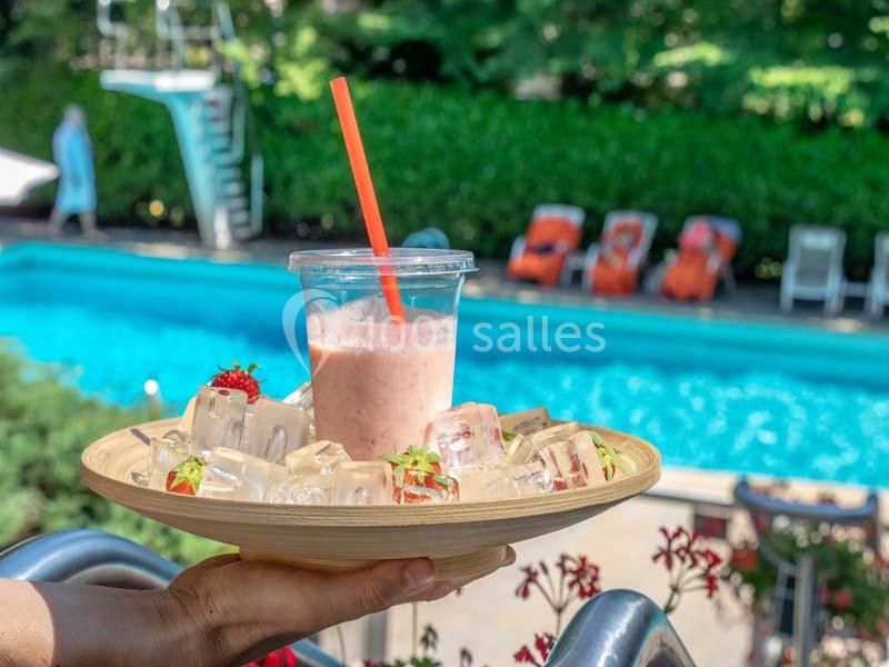 Un plateau avec des glaçons et des fraises entoure un smoothie dans un verre, près d'une piscine extérieure.