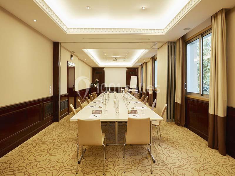 Salle de réunion lumineuse avec une grande table rectangulaire entourée de chaises, équipée d'un écran et d'un paperboard.