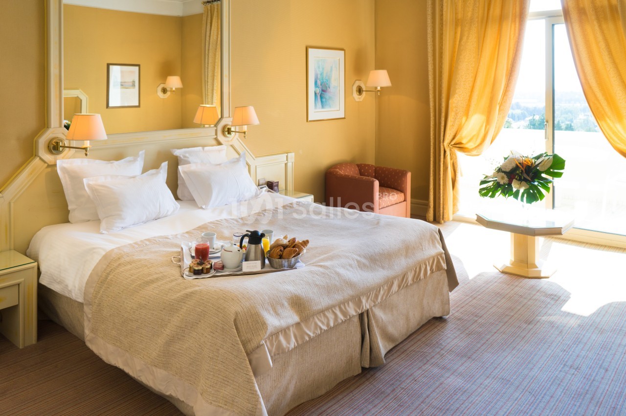 Chambre d'hôtel lumineuse avec lit double, plateau petit-déjeuner, fauteuil, table basse et grande fenêtre avec rideaux.