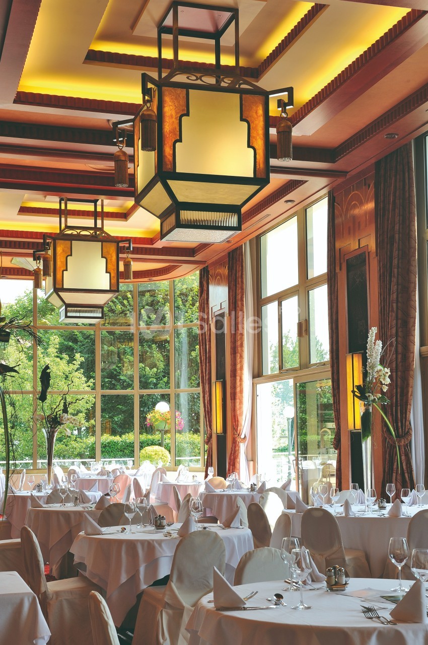 Salle de restaurant élégante avec grandes baies vitrées, tables dressées et luminaires suspendus au plafond.