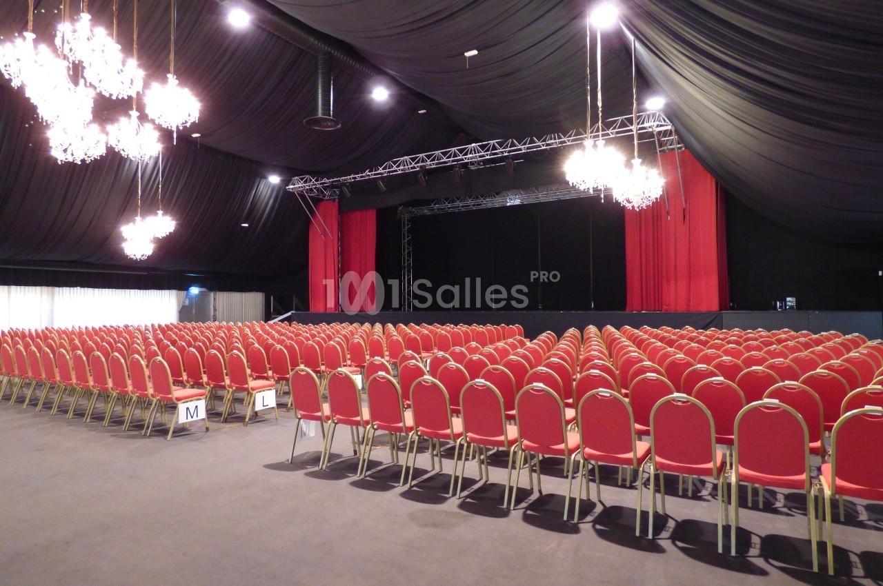 Salle de spectacle avec rangées de chaises rouges alignées face à une scène entourée de rideaux noirs et rouges.