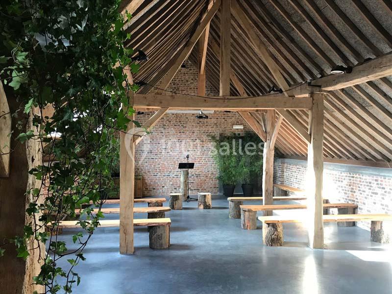 Salle en bois avec poutres apparentes, bancs rustiques en rondins et plantes vertes, éclairée par la lumière naturelle.