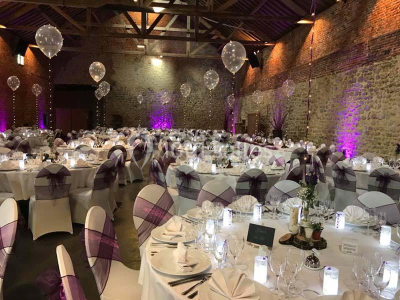Salle de réception décorée avec des tables rondes, nappes blanches, chaises ornées de rubans violets et ballons suspendus.