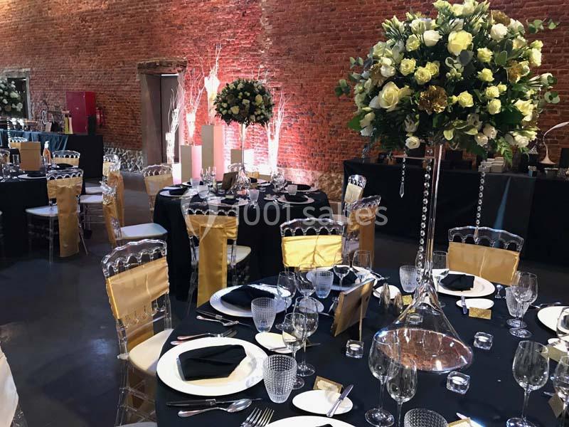 Salle de réception décorée avec des tables rondes, nappes noires, chaises transparentes et centres de table fleuris.