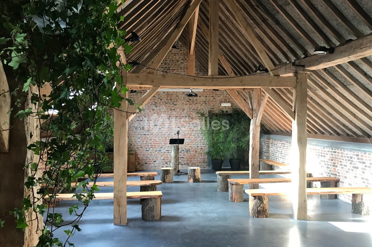 Salle avec charpente en bois apparente, bancs rustiques en rondins, murs en briques et plantes vertes décoratives.