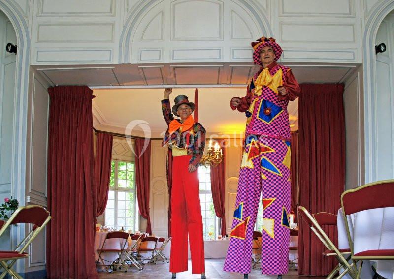 Deux artistes sur échasses en costumes colorés se tiennent dans une salle avec des rideaux rouges et des fenêtres.
