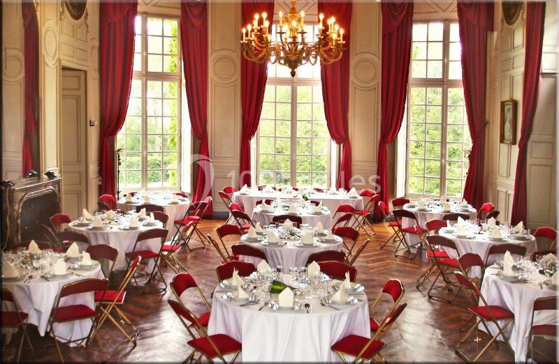 Salle élégante avec tables rondes dressées, chaises rouges, grandes fenêtres et rideaux rouges, lustre central.