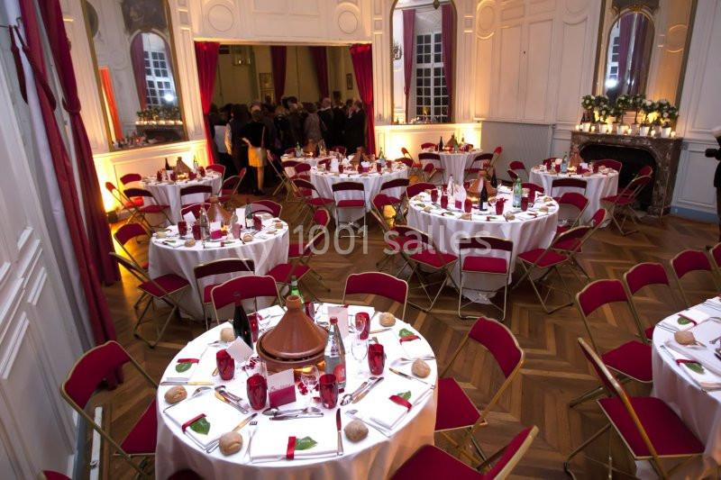 Salle élégante avec tables rondes dressées, nappes blanches, chaises rouges et lumière tamisée pour un événement.