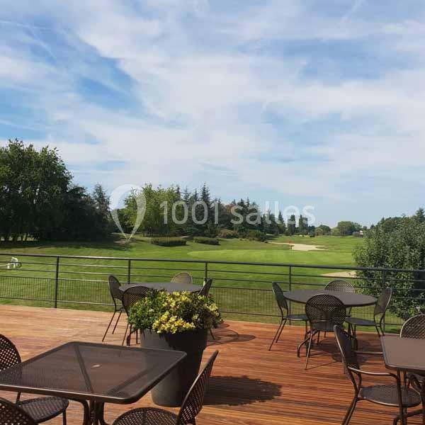 Terrasse en bois avec tables et chaises, surplombant un terrain de golf verdoyant sous un ciel partiellement nuageux.