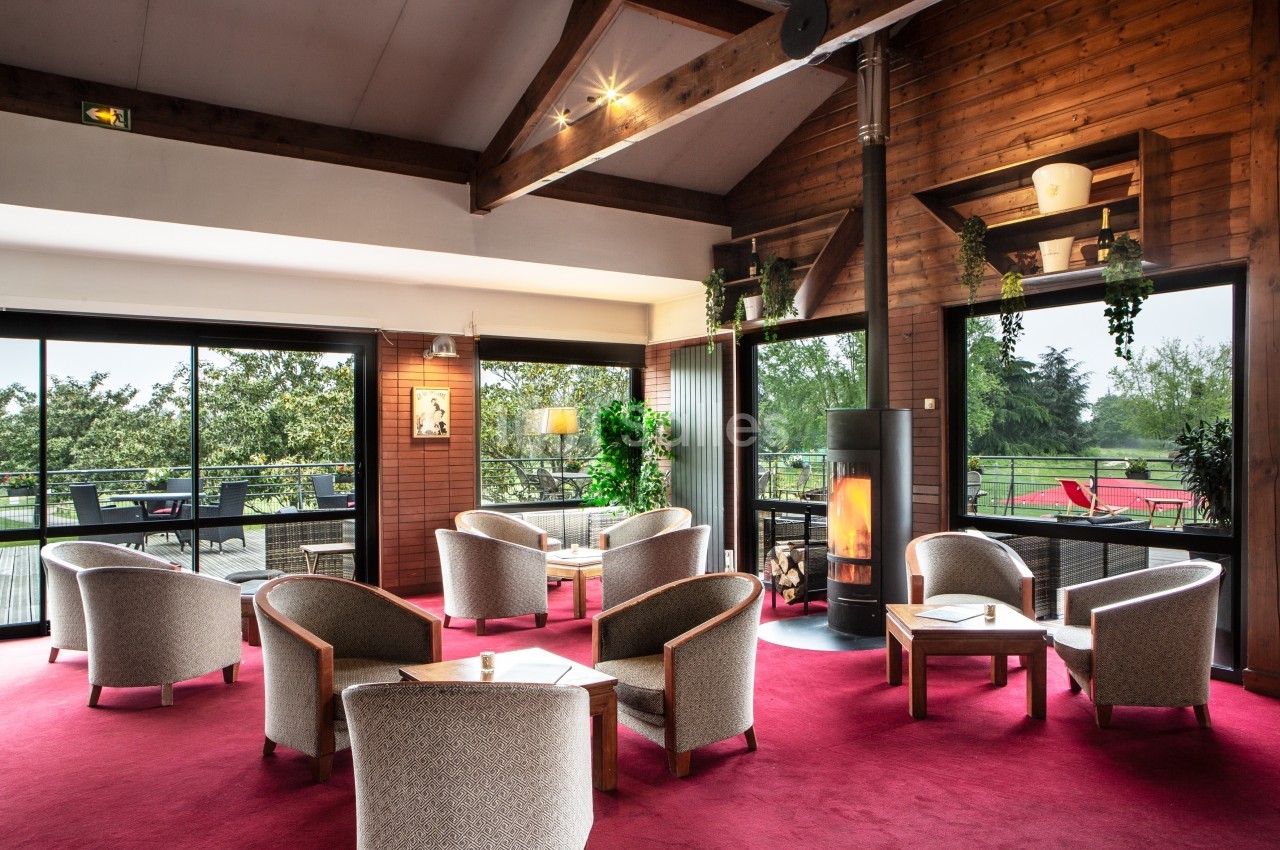 Salon chaleureux avec fauteuils, cheminée allumée, grandes baies vitrées et vue sur un espace extérieur verdoyant.