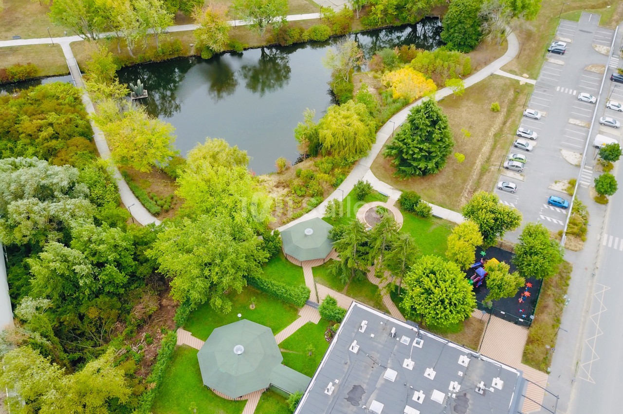 Vue aérienne d'un parc avec étang, sentiers, espaces verts, aire de jeux et parking adjacent.