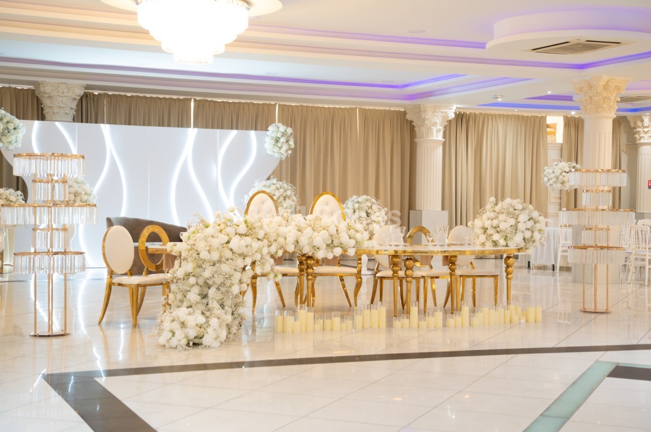 Salle de réception élégante avec décoration florale blanche, chaises dorées et éclairage tamisé.