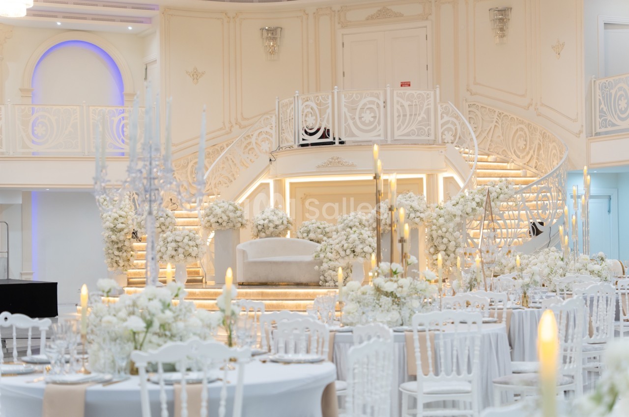 Salle de réception élégante avec décorations florales blanches, escalier central et tables dressées.
