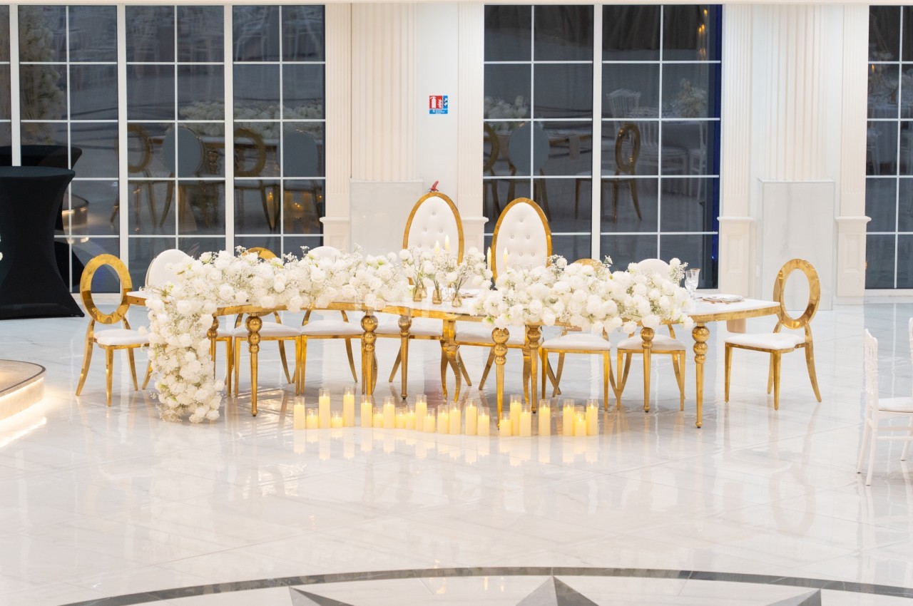 Table décorée avec des fleurs blanches, chaises dorées et bougies au sol dans une salle lumineuse avec grandes fenêtres.