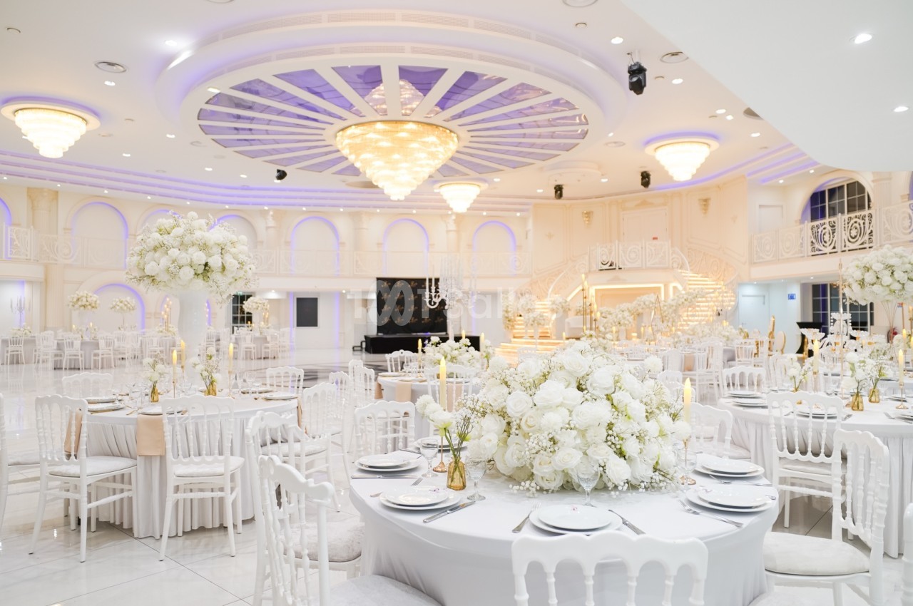 Salle de réception élégante avec tables décorées de fleurs blanches, chaises blanches et grand lustre central.